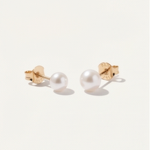 A pair of 5mm Dainty pearl stud earrings with gold posts, shown on a light cream background 