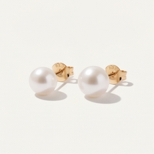 A pair of 7mm round pearl stud earrings with gold posts, shown on a light cream background 