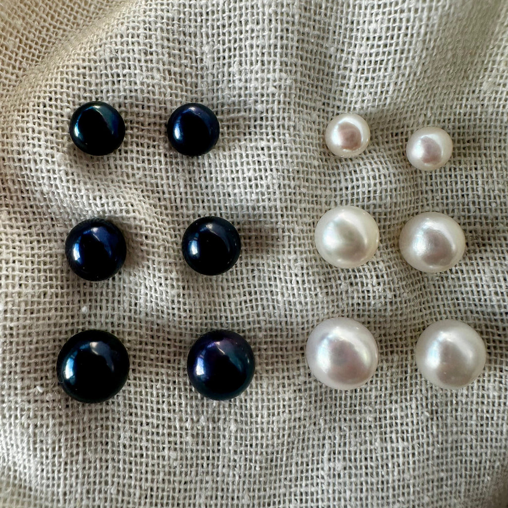 Six pairs of pearl-like stud earrings arranged on a textured beige fabric background, with four dark iridescent black pearls on the left and four white pearls on the right.