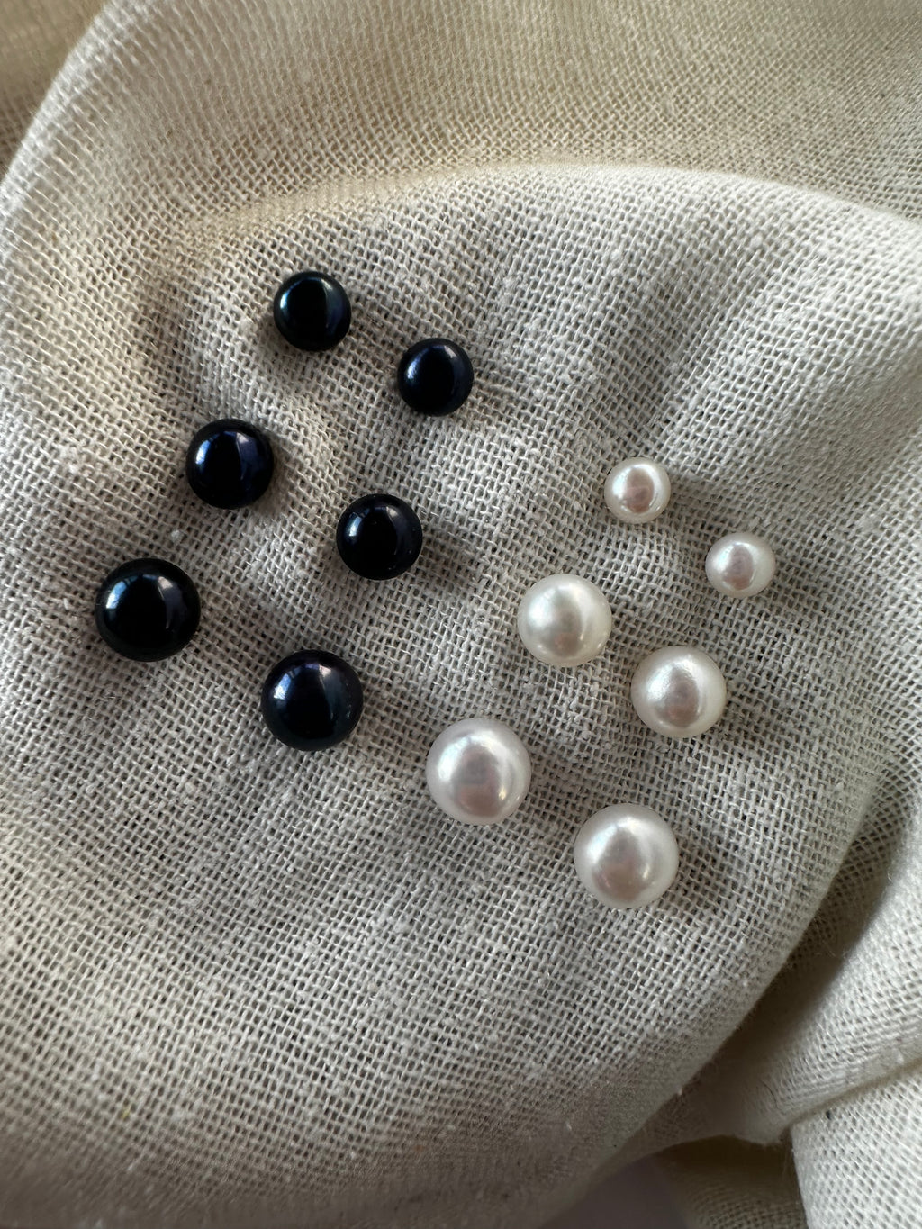 Nine round faux pearl beads, including seven black and two white pearls of varying sizes, arranged on a textured beige linen fabric background.