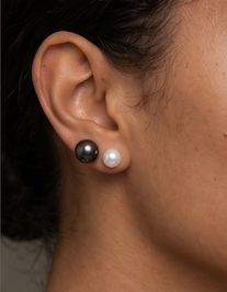 Close-up of a person's ear wearing two pearl stud earrings, one dark gray and one white, against dark hair and skin.