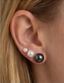 Three pearl stud earrings in white and black sizes worn on a person's ear, shown in a close-up lifestyle shot.