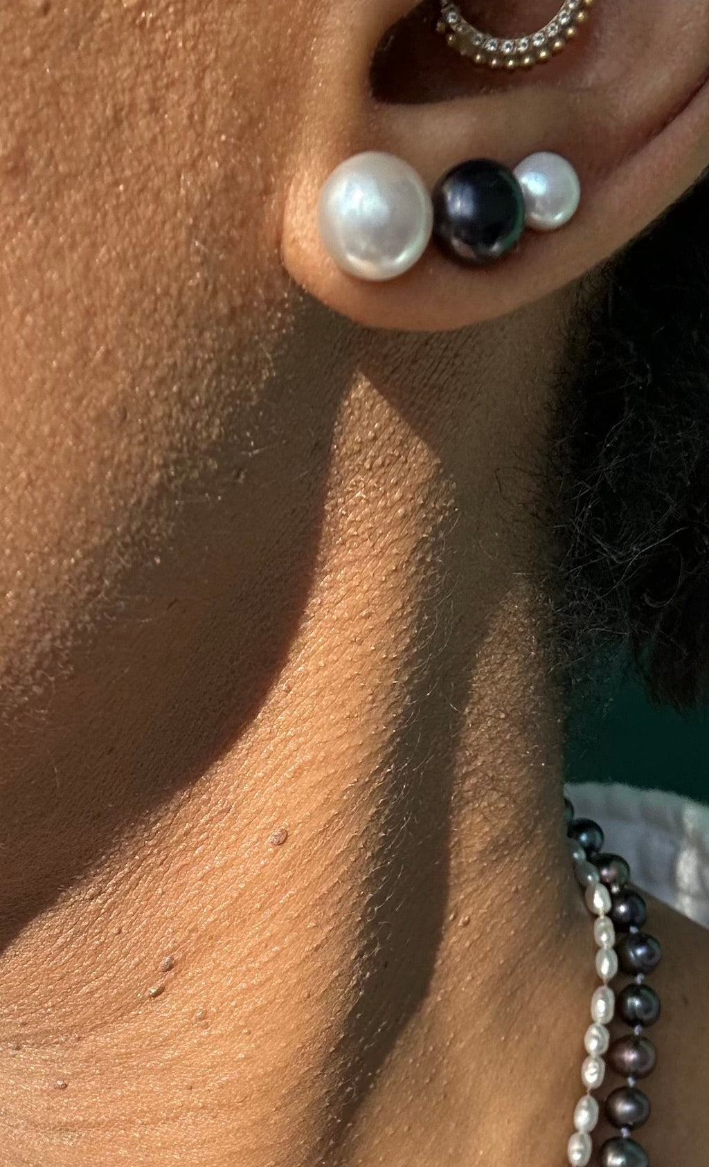 Close-up of a person wearing two beaded necklaces: one white pearl strand and one dark gray to black pearl strand, with matching stud earrings visible on the ear.