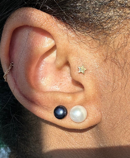 Close-up of a person's ear wearing multiple earrings, including a small gold star stud, a gold hoop, and a pair of round pearl-like studs in dark and white, with a pearl necklace visible below.